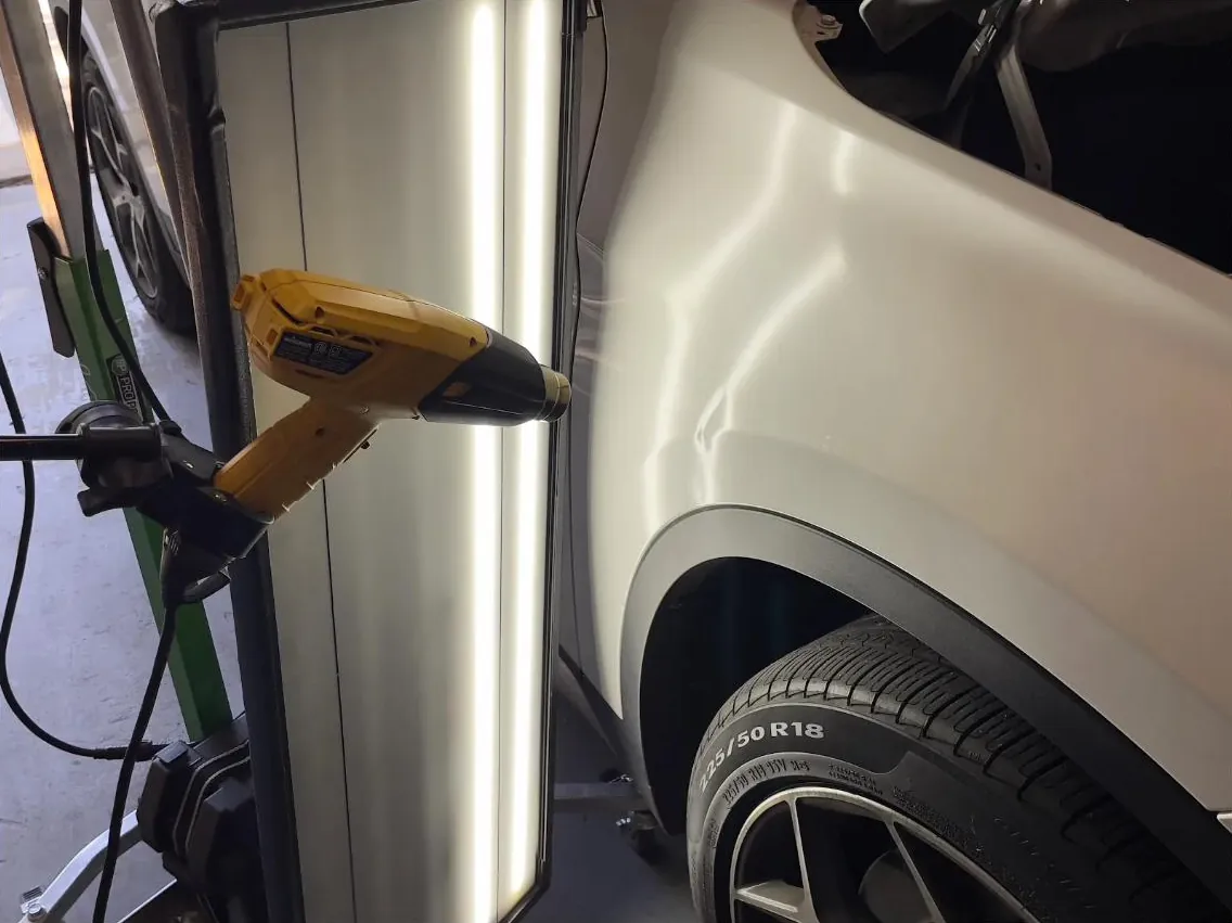 Technician warming a quarter panel with a heat gun beside the PDR light board